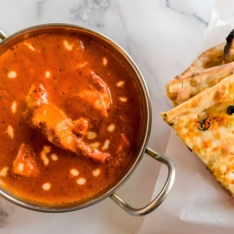 A metal pot of vibrant orange Indian butter chicken curry with naan bread on a white surface.