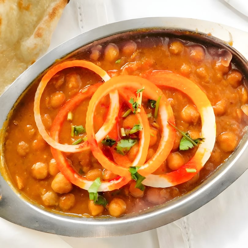 A metal bowl of Punjabi Dhaba Dixon's chana masala, garnished with onion rings and cilantro, with naan bread.