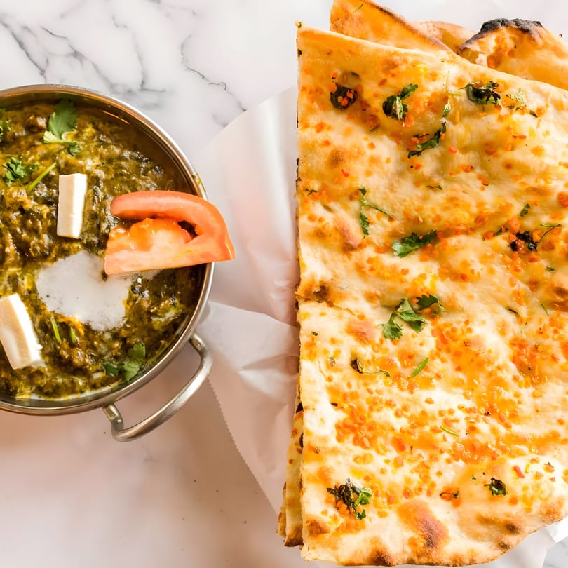 Palak Paneer with fresh paneer, tomato, and cilantro, served with garlic naan on marble.