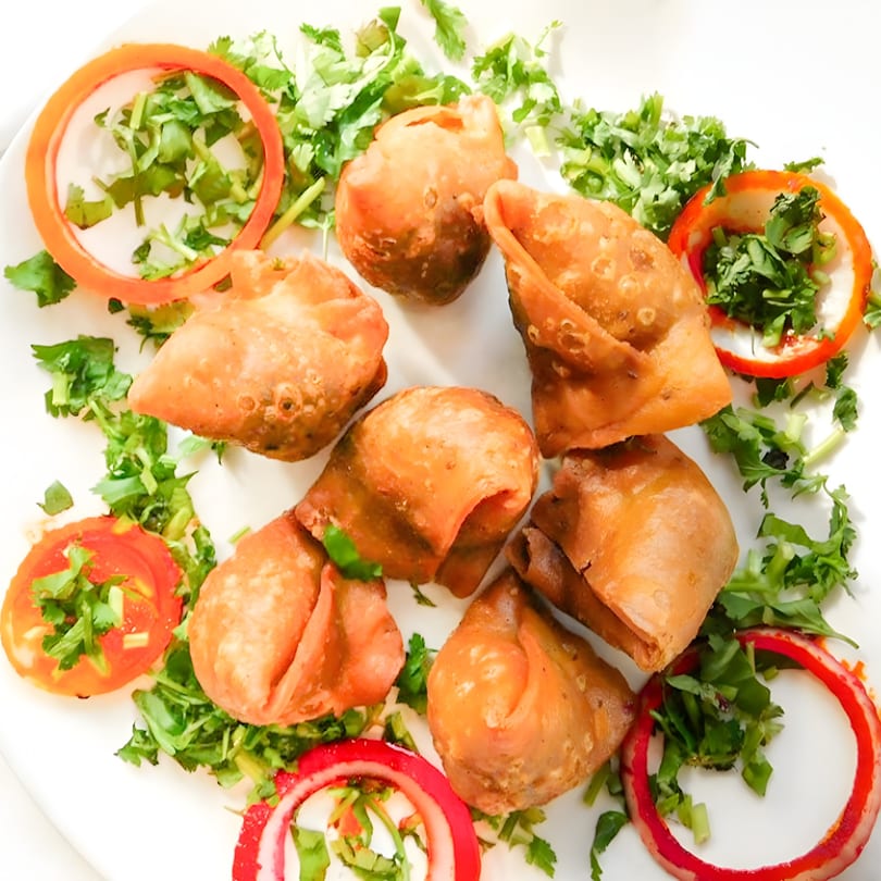 Seven golden-brown samosas garnished with fresh cilantro, tomato, and red onion slices on a white plate.