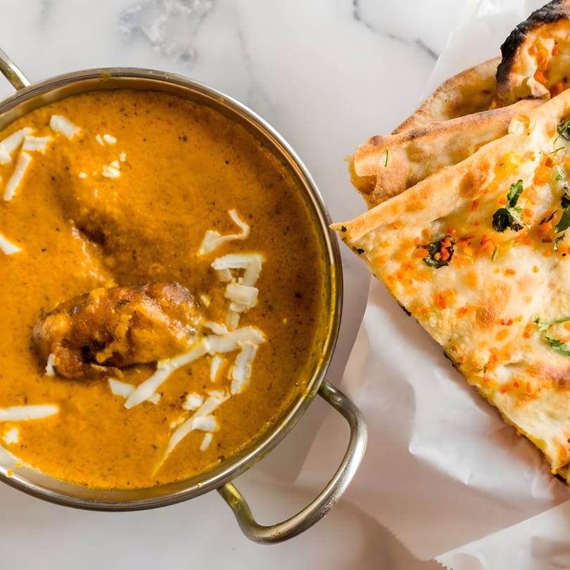 Indian butter chicken in a metal bowl with naan bread on the side, on a marble surface.